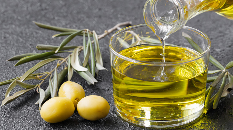 olive oil being poured into a small glass on a granite counter next to rosemary and green olives