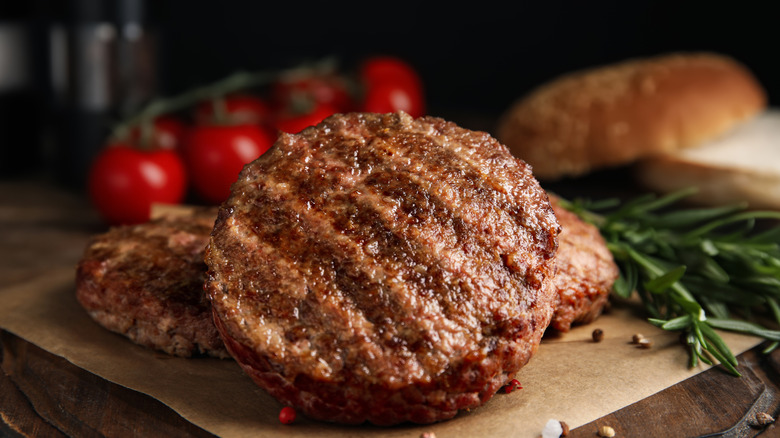 Grilled hamburger patty leaning on two others on brown paper with round tomatoes, a bun, and herbs surrounding it