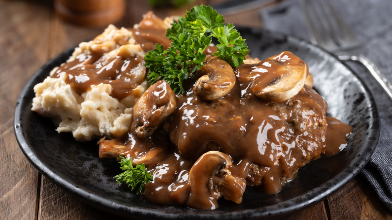 Salisbury steak covered with brown mushroom gravy on a black plate with gravy-covered mashed potatoes and a parsley garnish