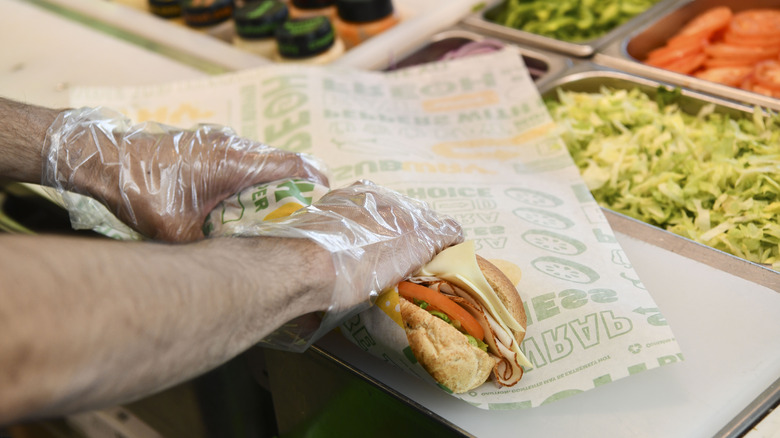 A Subway employee wearing plastic gloves wraps a footlong sandwich behind a counter