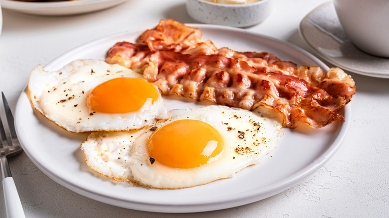 A white plate is filled by a pair of fried eggs and two strips of bacon