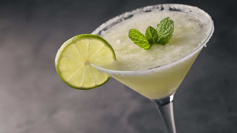 A daiquiri in a martini glass garnished with a lime, mint leaves, and a salt rim