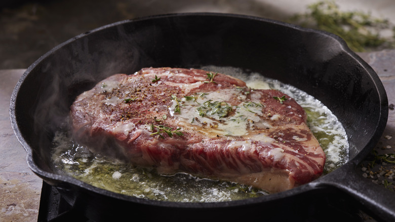 A steak topped with herbs and butter cooking in a black cast iron pan