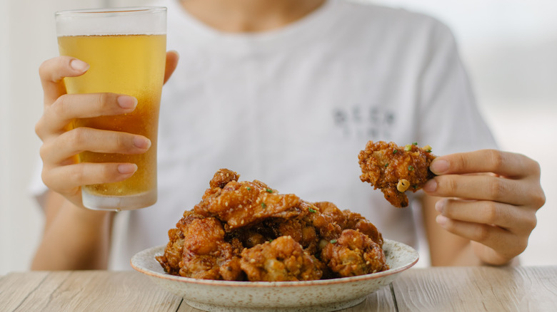 Person eating Korean fried chicken and holding a glass of beer