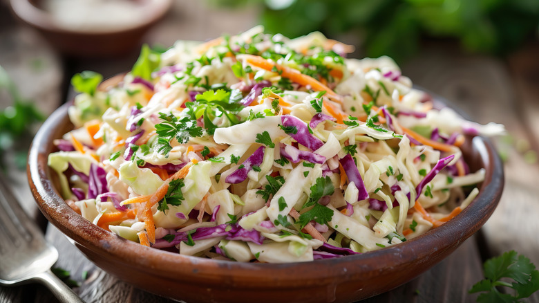 Bowl of coleslaw with fork next to it and blurry greens and small bowl of sauce in the background