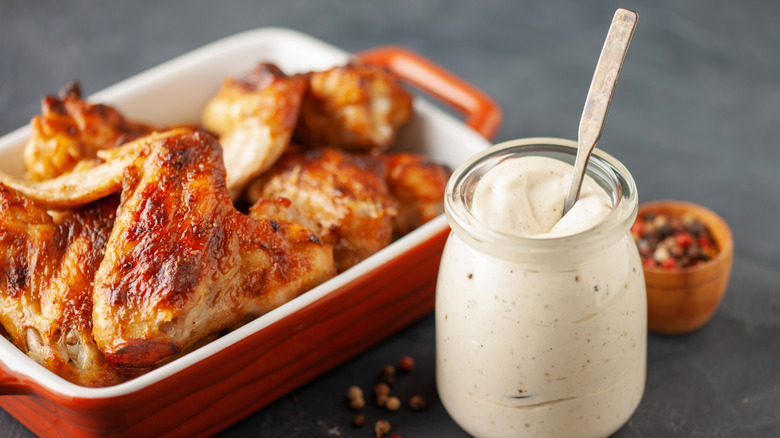 Small jar of Alabama white sauce with spoon in it with dish of chicken wings and small container of peppercorns