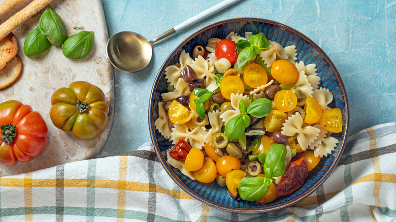 A bowl of pasta salad topped with sliced cherry tomatoes, sun dried tomatoes, olives, and fresh basil, surrounded by fresh whole tomatoes, basil, and bread sticks.