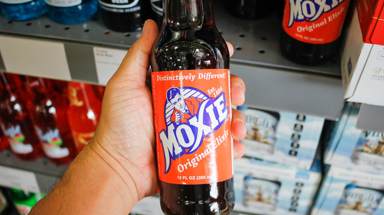 Hand holding a bottle of Moxie soda in front of store shelves