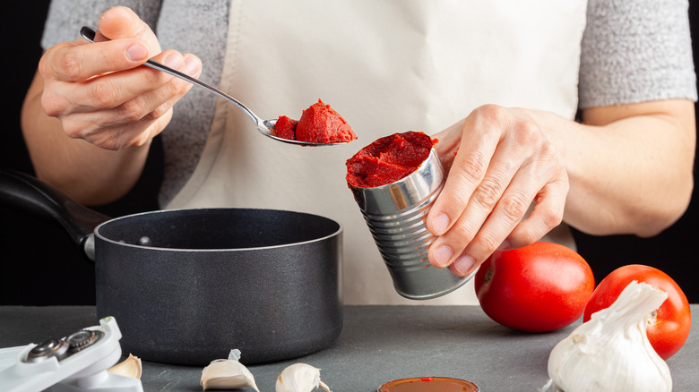 A person adding a spoonful of tomato paste from an open can into a pot for mixing with other ingredients