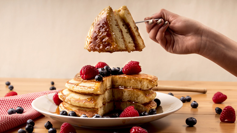 A fork holds a triangular bite of food above a stack of fluffy pancakes with berries.