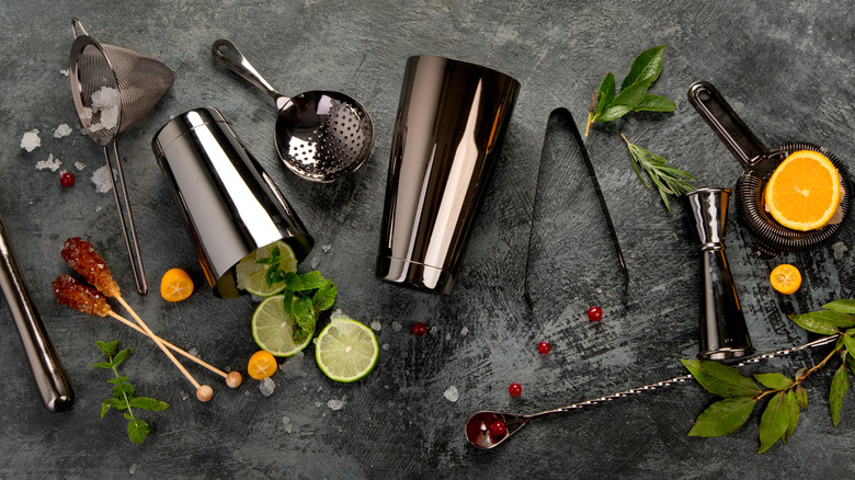 Assorted metal bar tools with slices of citrus fruit, herbs, and berries on a gray slate surface