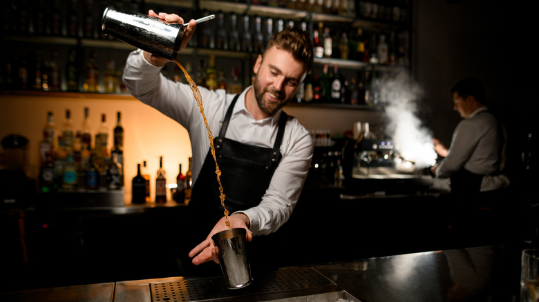 Bartender behind a bar holding a metal shaker tin high and pouring a long stream of a drink into another shaker tin below