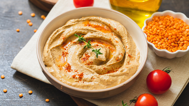 A bowl of red lentil hummus beside a bowl of red lentils on a napkin with cherry tomatoes