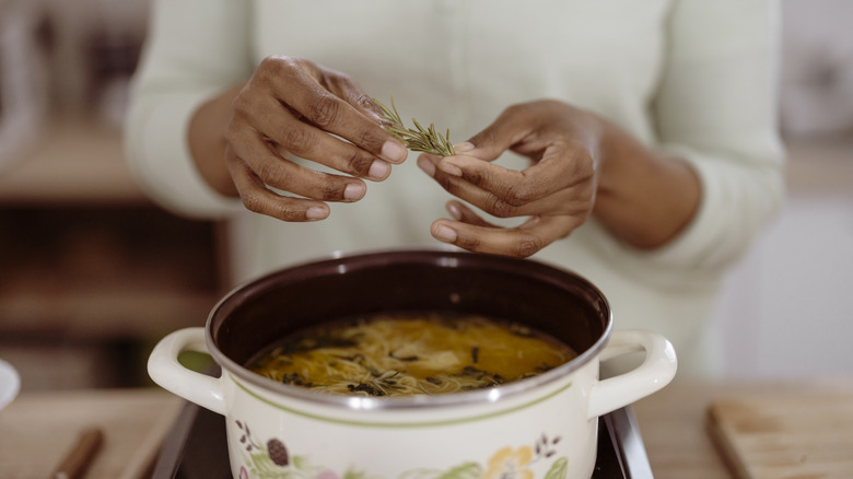 Hands holding herb sprig over pot of soup