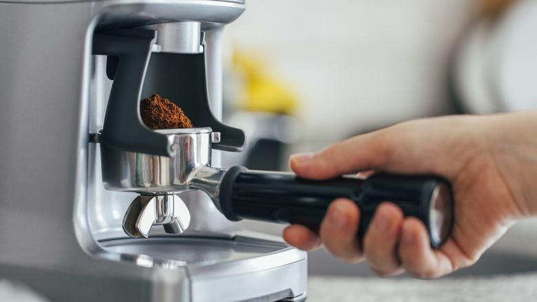 Hand holding group handle heaped with coffee grounds under coffee grinder