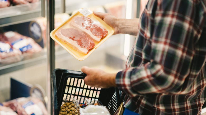 Close up of a man shopping for raw, red meat while holding a shopping basket