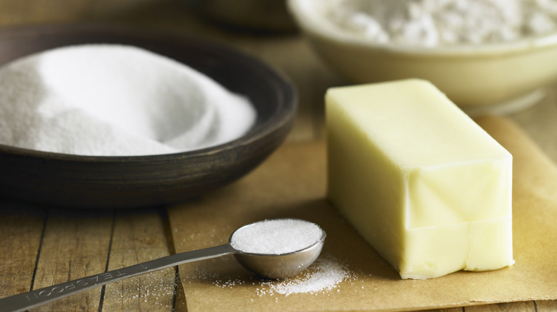 Stick of butter on parchment paper next to bowl and spoon full of white sugar