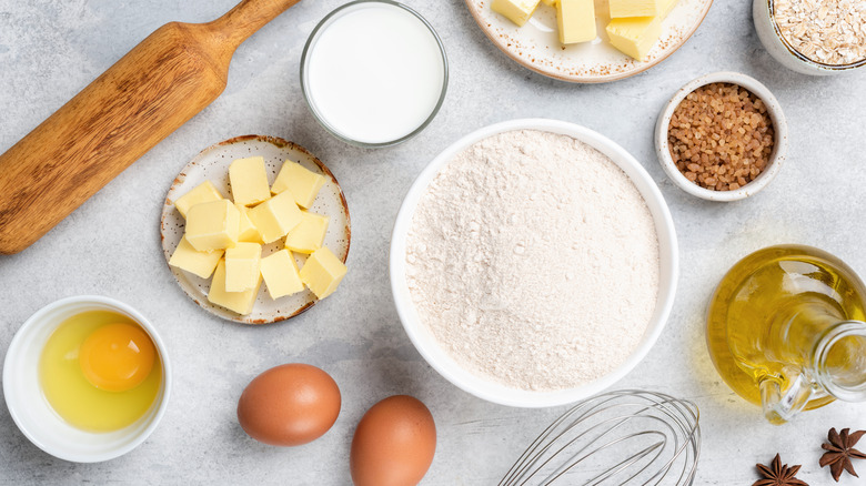 A wide variety of baking ingredients in bowls including eggs, flour, and butter on gray surface