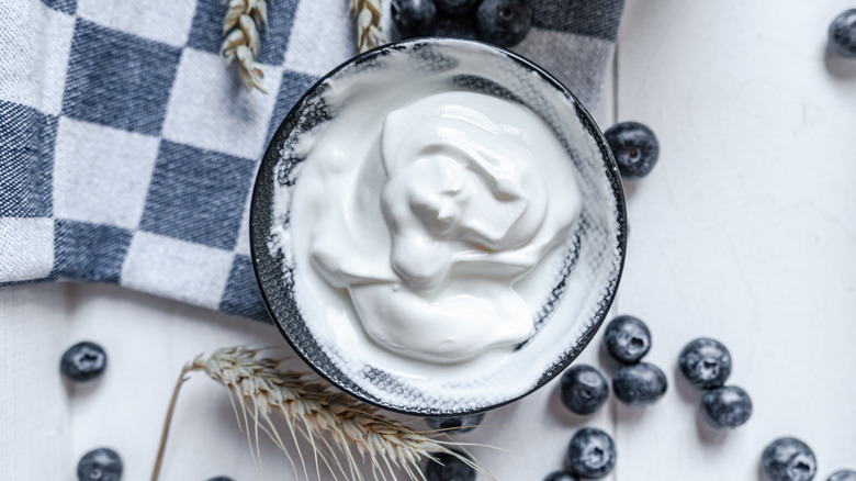 Top view of a bowl of greek yogurt on a checkered cloth, surrounded by blueberries and wheat