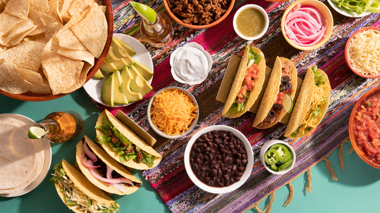 A selection of tacos sit on a table next to several condiments, tortilla chips, and beer bottles with lime wedges