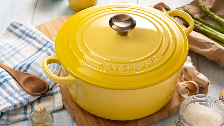 A yellow Dutch oven made by Le Creuset is surrounded by various ingredients, including garlic and asparagus