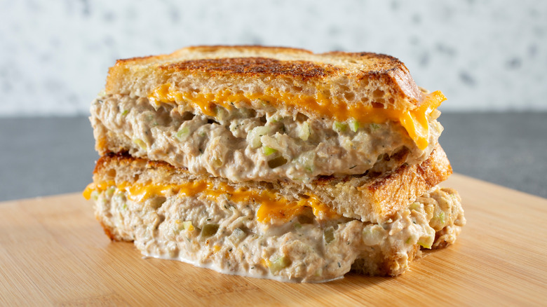 A stacked tuna melt has melty cheese running down its sides and creamy tuna salad dripping out onto a wooden cutting board