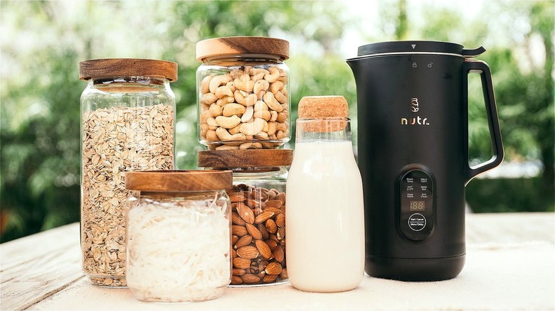 Black Nutr nut milk machine next to glass jars of milk, nuts, and oats