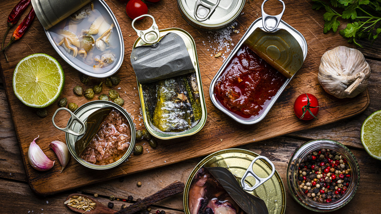 A chopping board filled with open tins of fish and ingredients surrounding the tins such as cherry tomatoes, half a lime, garlic cloves, and peppers