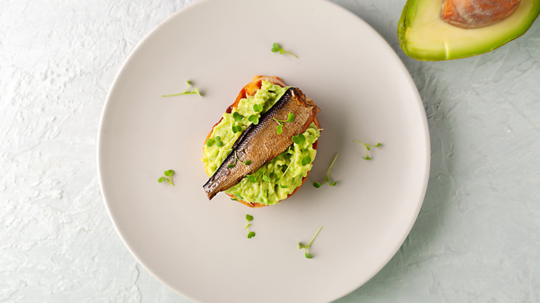 A plate with one slice of avocado toast topped with canned fish and watercress and half an avocado on the side