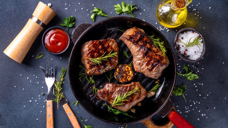 Three cooked steaks in a pan with fresh herbs, olive oil, salt, and pepper