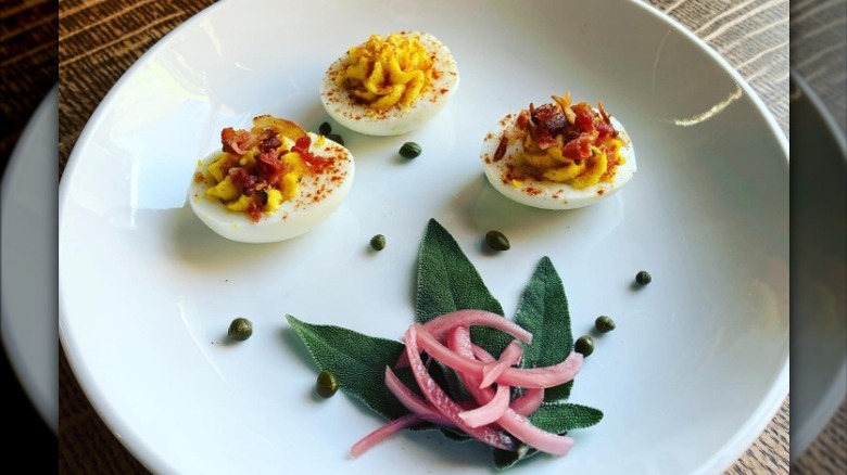 A plate of devilled eggs is topped with bacon crumbles, paprika, and is scattered with capers, pickled onion, and sage leaves