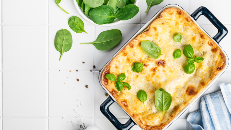 Pan with white cheese topped lasagna and basil leaves on top and next to the pan on a white surface