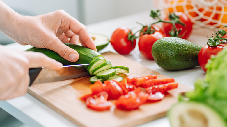 Someone's cutting a cucumber next to a pile of sliced tomatoes on a cutting board.