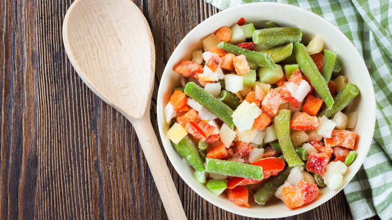 White bowl full of frozen vegetables including green beans, cauliflower, and carrots next to white wooden spoon