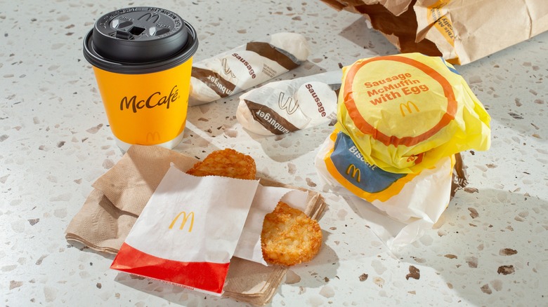 A spread of McDonald's breakfast items including a coffee cup, two hashbrowns, wrapped sausage McMuffins, and breakfast burritos