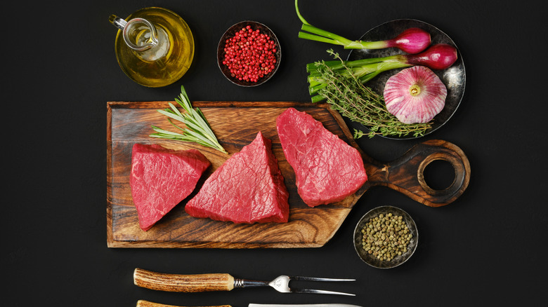 Three small pieces of beef on a small wooden cutting board on a black surface, surrounded by small bottle of oil, small bowls of herbs and spices, and wooden-handled fork