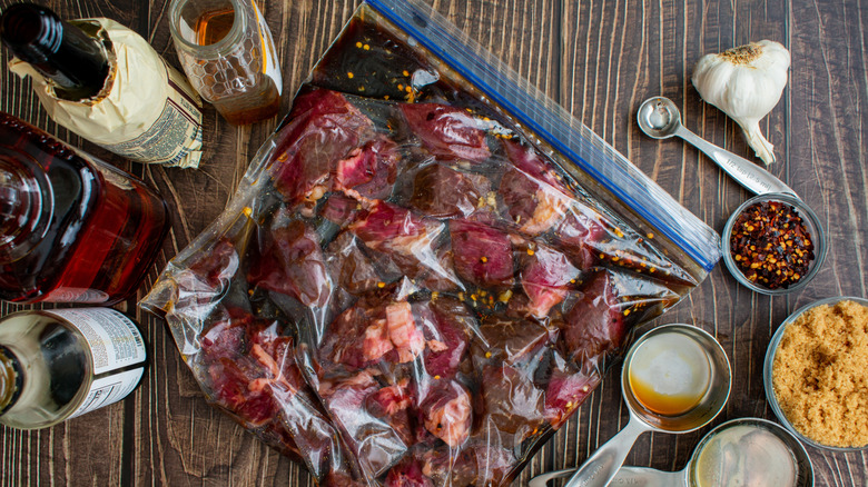 Steak tips being marinated in a plastic bag on a wood surface surrounded by garlic small bowls of ingredients, small ingredient bottles, and metal measuring cups