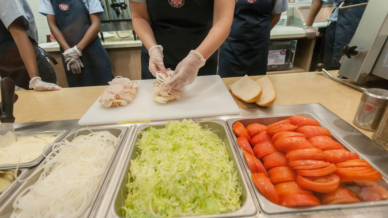 Long metal containers of sliced onions, tomatoes and shredded lettuce in front of a Jersey Mike's worker making a sub with other workers in the background