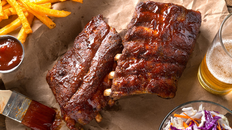 A plate of pork ribs on a platter, smothered in bbq sauce, surrounded by French fries, a sauce basting brush, a beer and coleslaw.