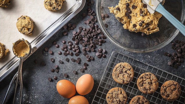 Freshly baked cookies on a wire rack next to a bowl of cookie dough, chocolate chips, and whole raw eggs