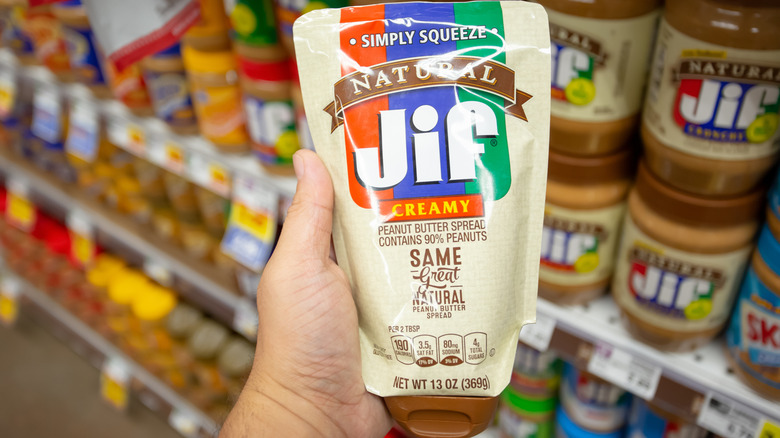 A hand holds a squeezable Jif peanut butter pouch in a grocery store peanut butter aisle