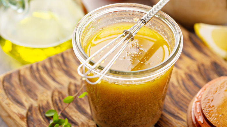 a homemade vinaigrette in glass jar on a wooden counter with small whisk balanced on top