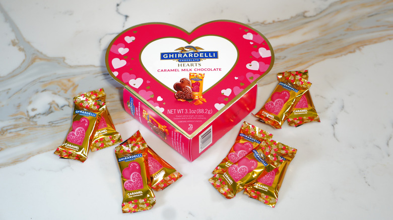 A closed heart-shaped Ghirardelli box with nine pieces of chocolate in wrappers surrounding the box