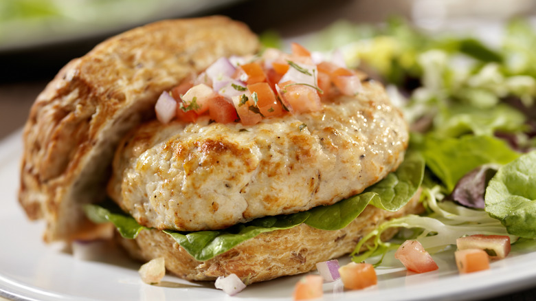 Coooked turkey burger patty with tomato and onion salsa on top on an open bun on a white plate with salad greens to the side