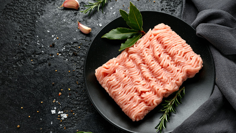 A rectangle of raw ground turkey on a black plate on a black surface with herb springs and garlic cloves on the side