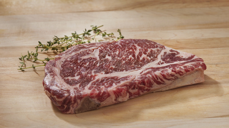 Raw ribeye with a lot of marbling sits next to thyme on a wooden table