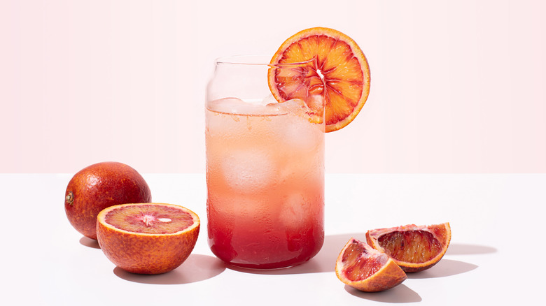 A blood orange cocktail with ice sits in a pink surface with a pink background surrounded by sliced and whole blood oranges