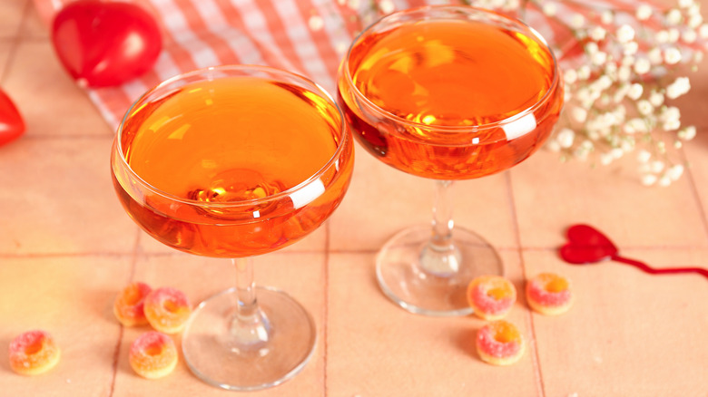 Two reddish cocktails in coupe glasses on a counter surrounded by hearts and candy