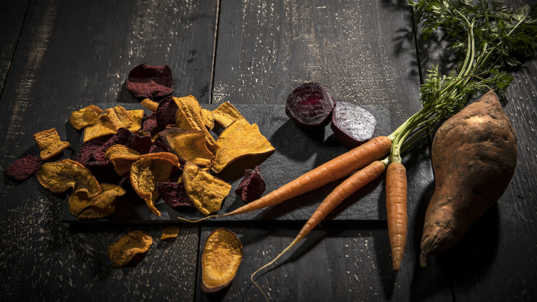 An array of vegetable chips sit on a wooden table next to carrots, sliced beets, and a sweet potato