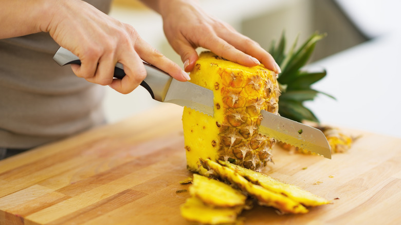 Woman's hands with serrated knife chopping a whole pineapple on cutting board